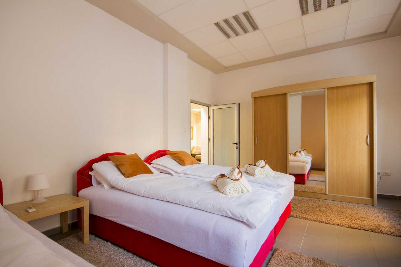 Two joined single beds with red bases and brown accent pillows, rolled towels with brown ribbons, large wood sliding wardrobe with mirrored door, and nightstand – Apartment Klio – City Break Apartments