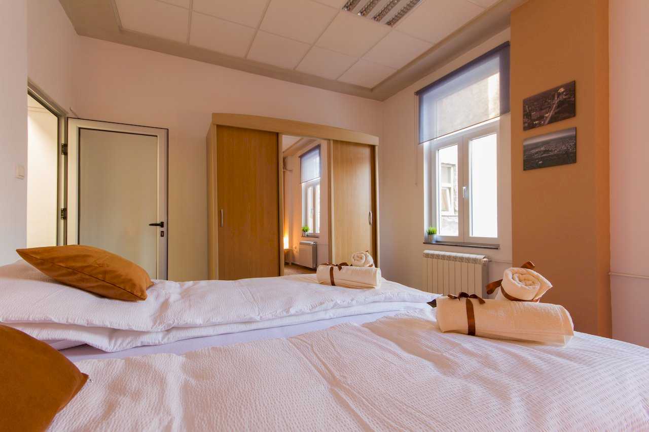 Bedroom with two beds, towels with brown ribbons, large wood wardrobe with full-length mirror on the center door, window with grey blind, and terracotta accent wall with two small framed pictures – Apartment Klio – City Break Apartments