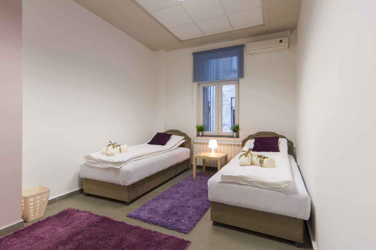 Two parallel single beds with brown bases and purple pillows, rolled towels with gold ribbons, shared nightstand with lamp, and blue roller blind on the window – Apartment Klio – City Break Apartments