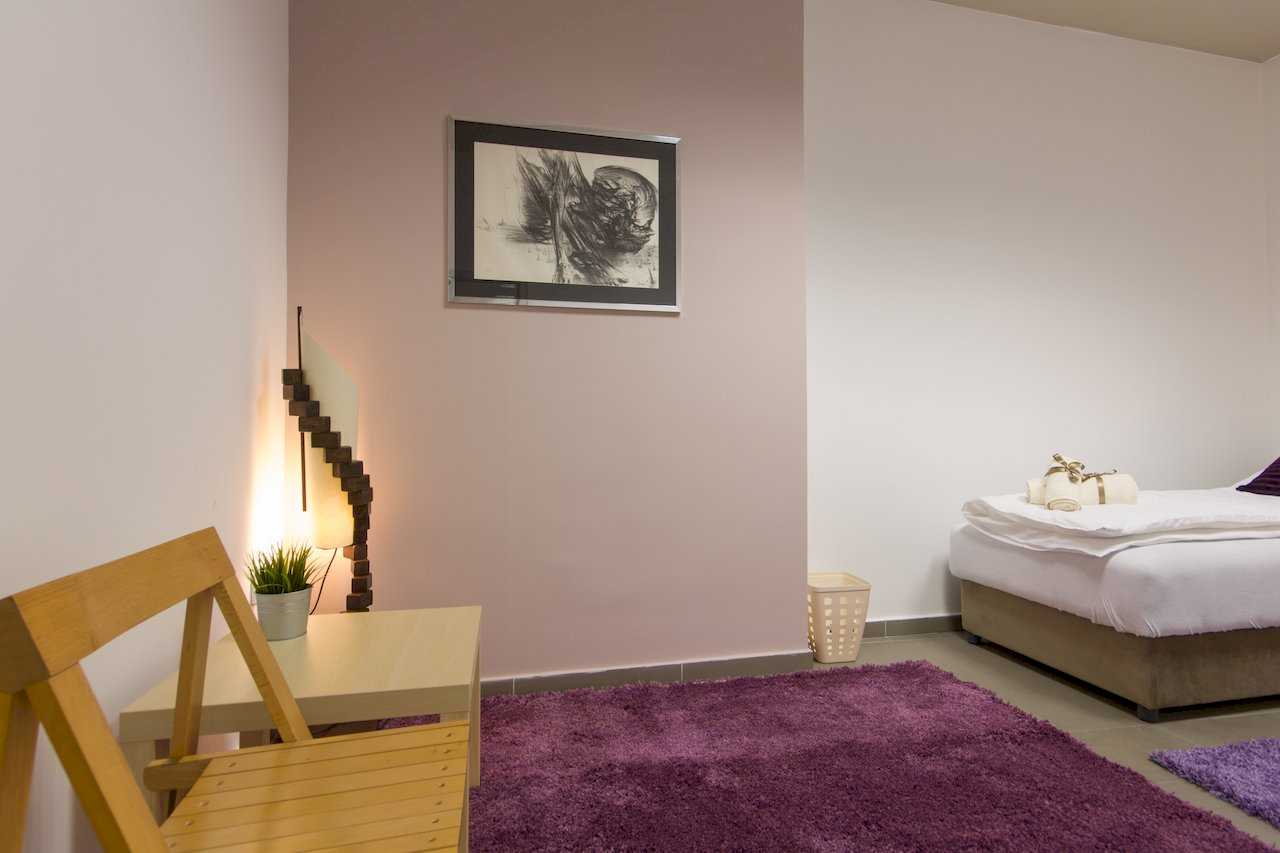 Bedroom with dusty-rose wall, abstract framed art, floor lamp, small wooden table and chair, purple shag rug – Apartment Klio – City Break Apartments