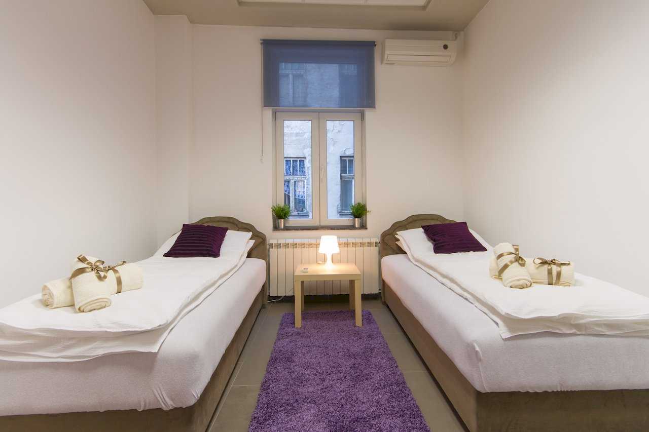Two single beds with beige frames and purple pillows, gold ribbons on rolled towels, purple shag rug between the beds, and a nightstand with lamp under the window – Apartment Klio – City Break Apartments