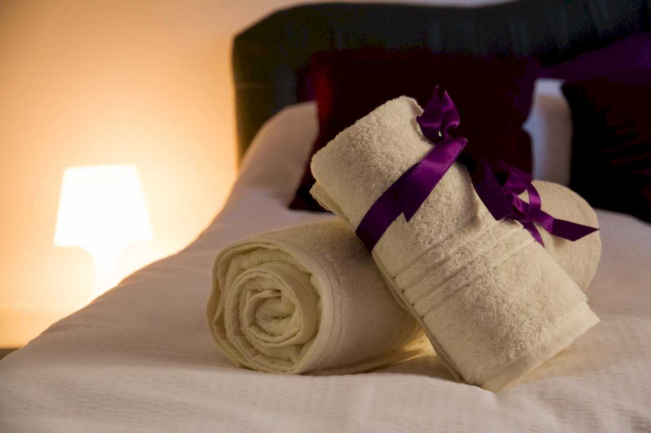 Close-up of rolled white towels with a purple satin ribbon on white bedding – Apartment Klio – City Break Apartments
