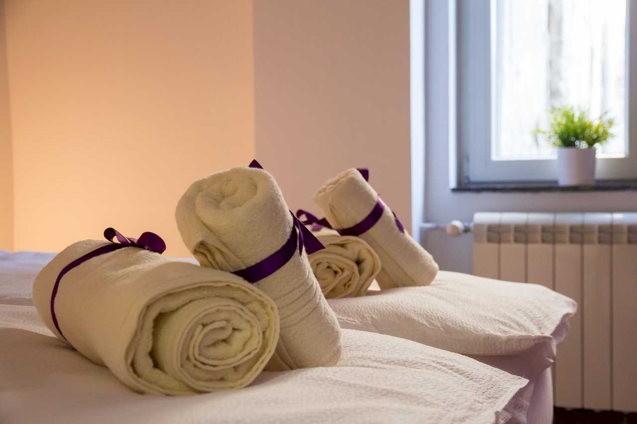 Close-up of four cream rolled towels tied with purple ribbons on white bedding – Apartment Klio – City Break Apartments