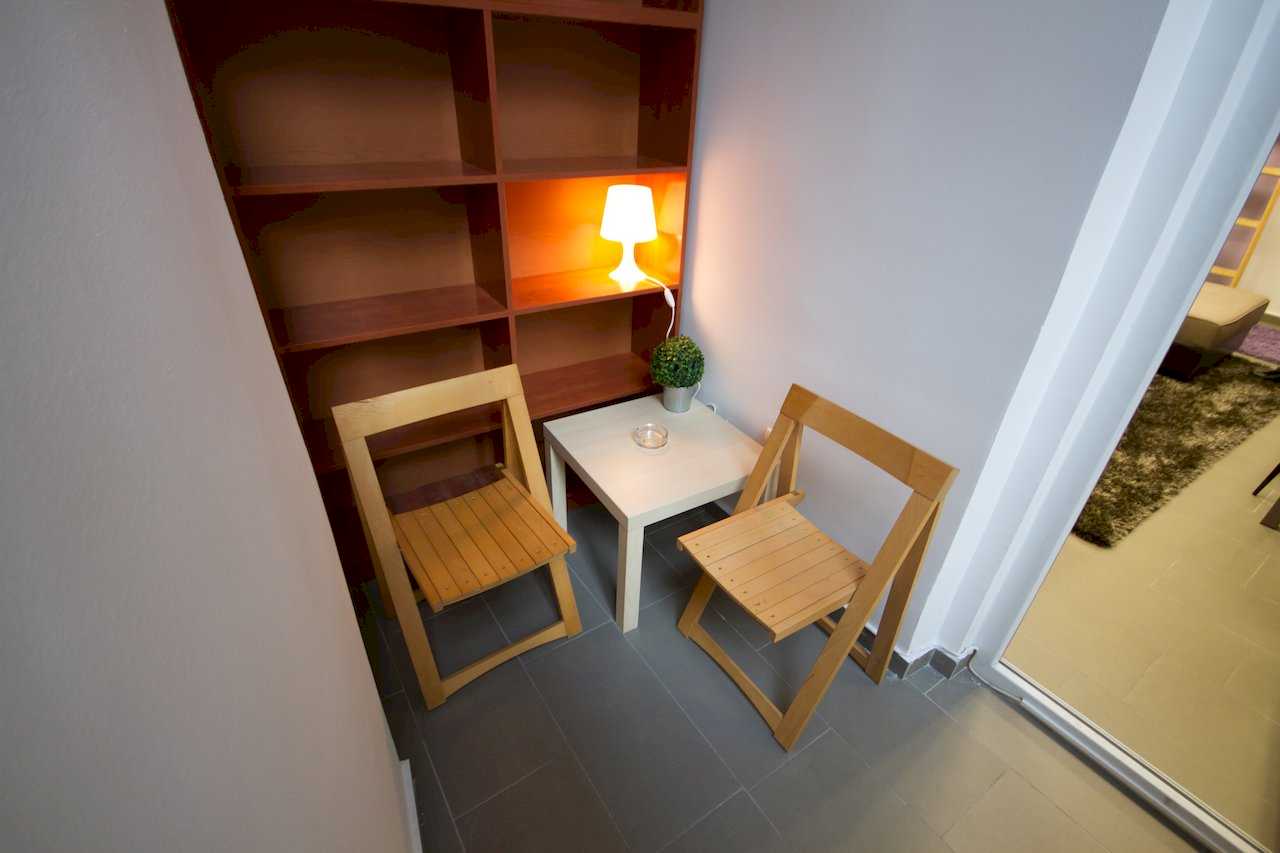 Small nook with two wooden folding chairs, white side table, built-in wood shelving unit, and a lit table lamp on a shelf – Apartment Klio – City Break Apartments
