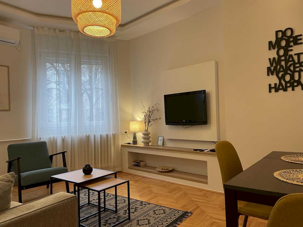 Living room with TV on a white panel, teal armchair, nesting tables, woven pendant light, partial dining set with mustard chairs and sheer curtained window – Klishe Zen Apartment – City Break Apartments