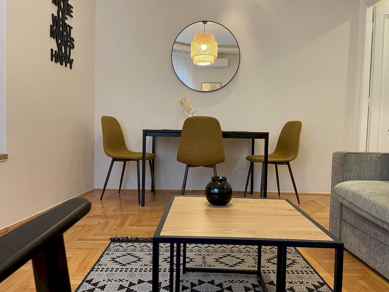Dining nook with black table, three mustard-yellow chairs, round mirror, typographic wall art, square coffee table and black-and-white geometric rug – Klishe Zen Apartment – City Break Apartments