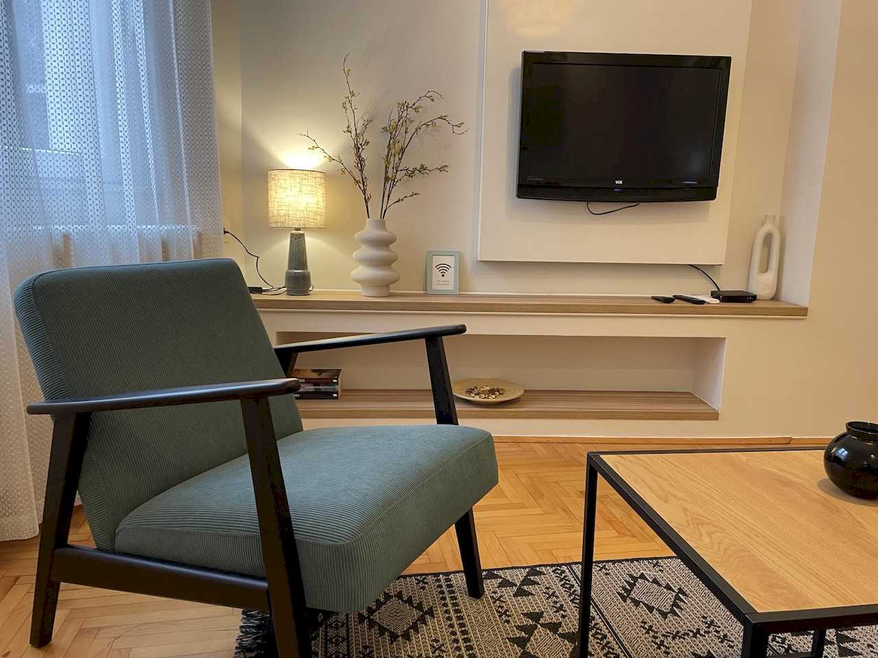 Living area with teal corduroy armchair, wall-mounted TV on white panel and built-in wood shelves with lamp, vase and Wi-Fi sign – Klishe Zen Apartment – City Break Apartments