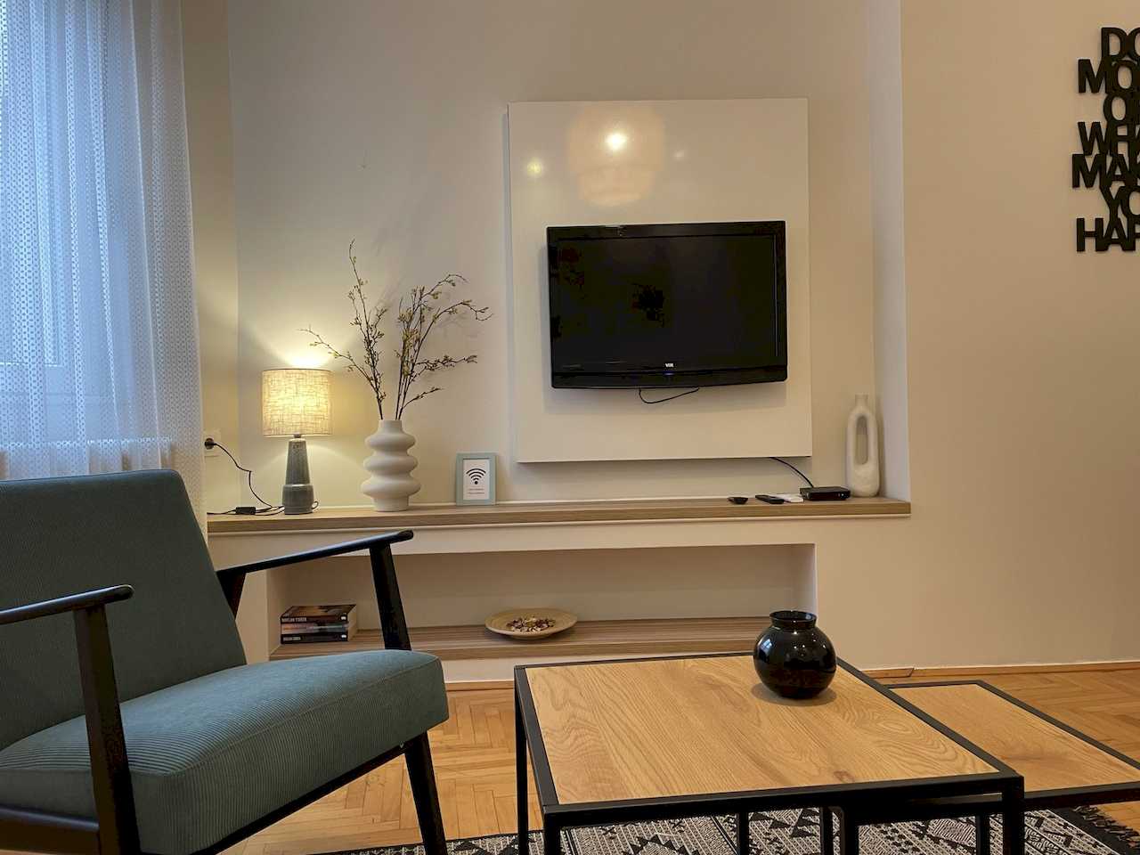 TV wall with white panel and wood shelf, teal armchair, nesting coffee tables and partial black typographic wall art – Klishe Zen Apartment – City Break Apartments