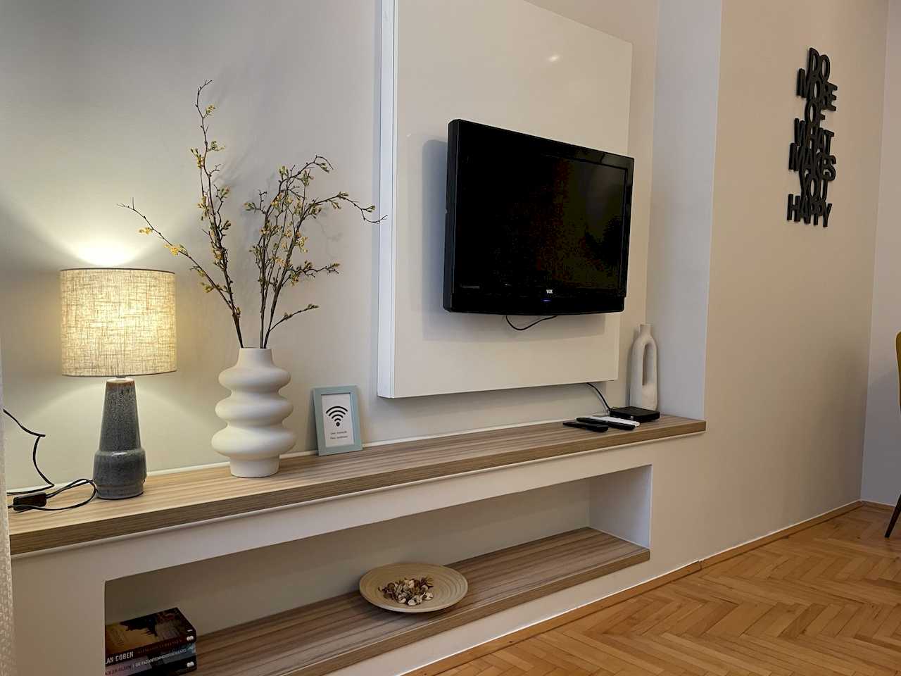 Close-up of the TV nook with wood shelves in a niche, table lamp, white vase with dried branches, Wi-Fi frame and wall quote – Klishe Zen Apartment – City Break Apartments