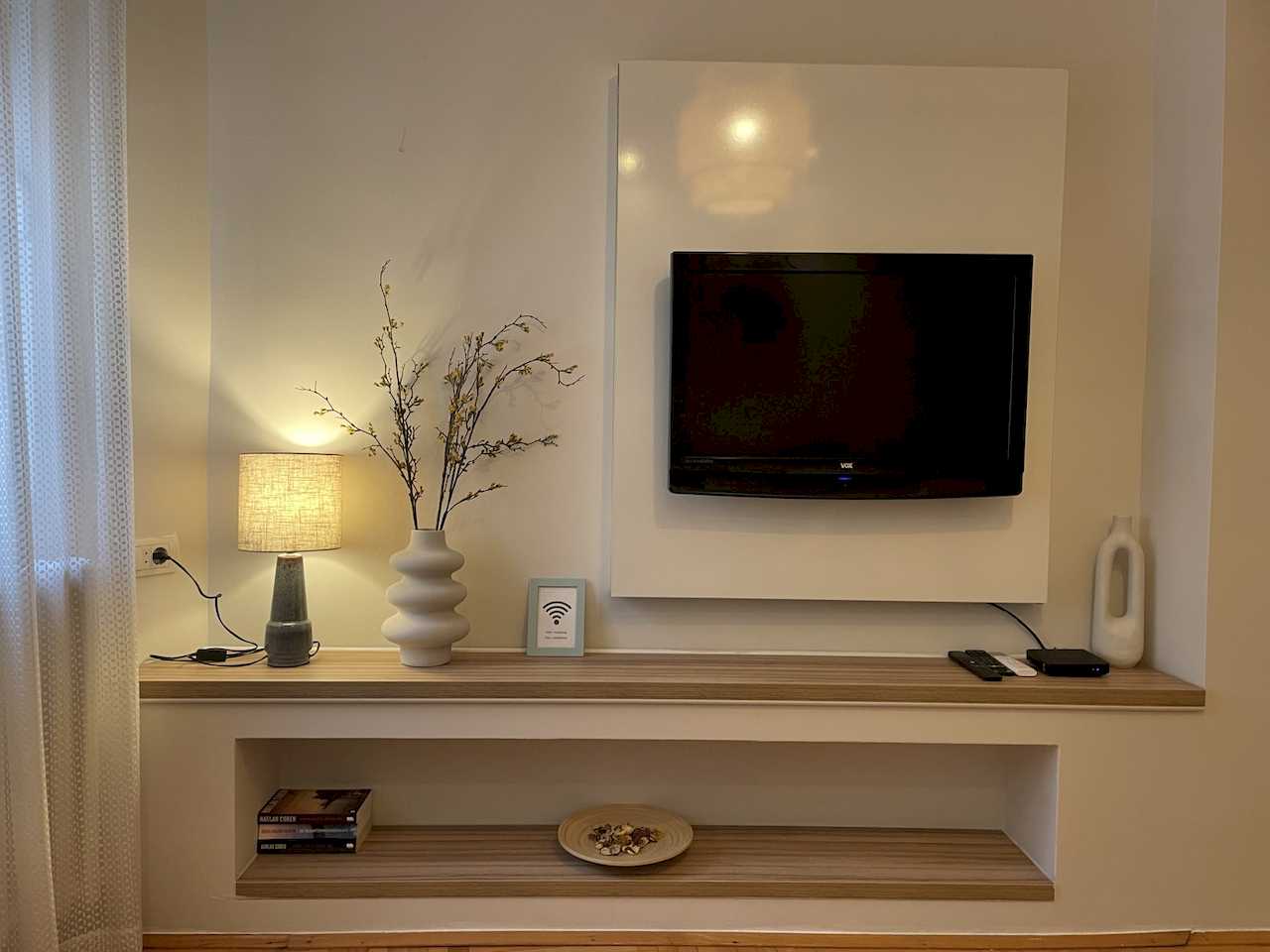 Wall-mounted TV on white panel with double wood shelf, lamp, vases, books and decorative bowl in the niche – Klishe Zen Apartment – City Break Apartments