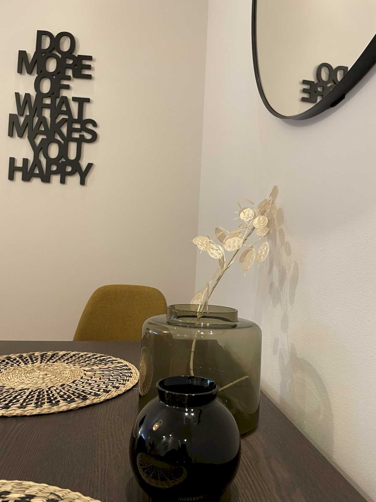 Dining table with woven placemats, glass vase with dried branches, black vase and black 3D wall quote – Klishe Zen Apartment – City Break Apartments