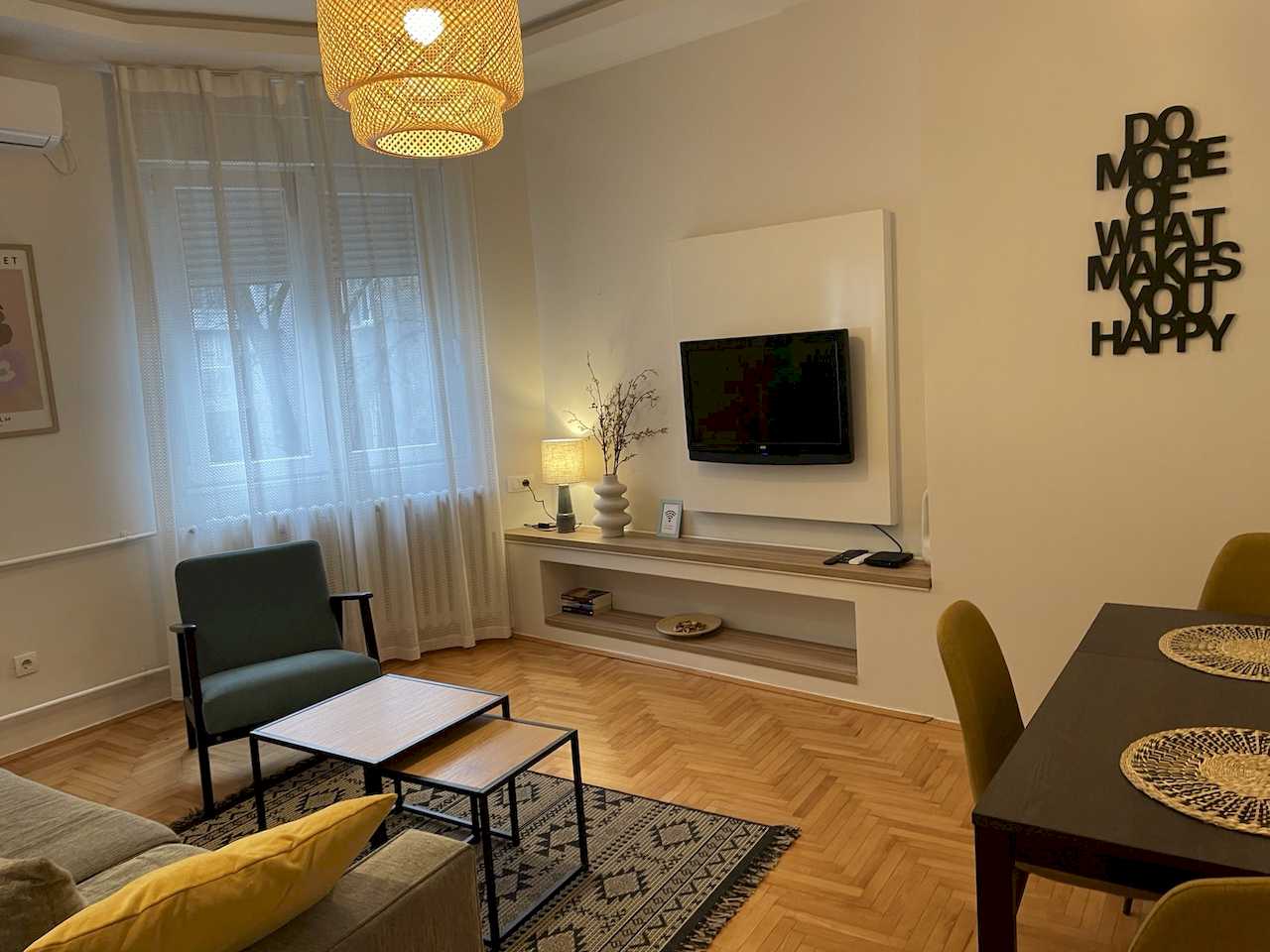 Open-plan living and dining – grey sofa with yellow cushion, teal armchair, nesting tables, woven pendant, TV area, mustard chairs and wall quote – Klishe Zen Apartment – City Break Apartments