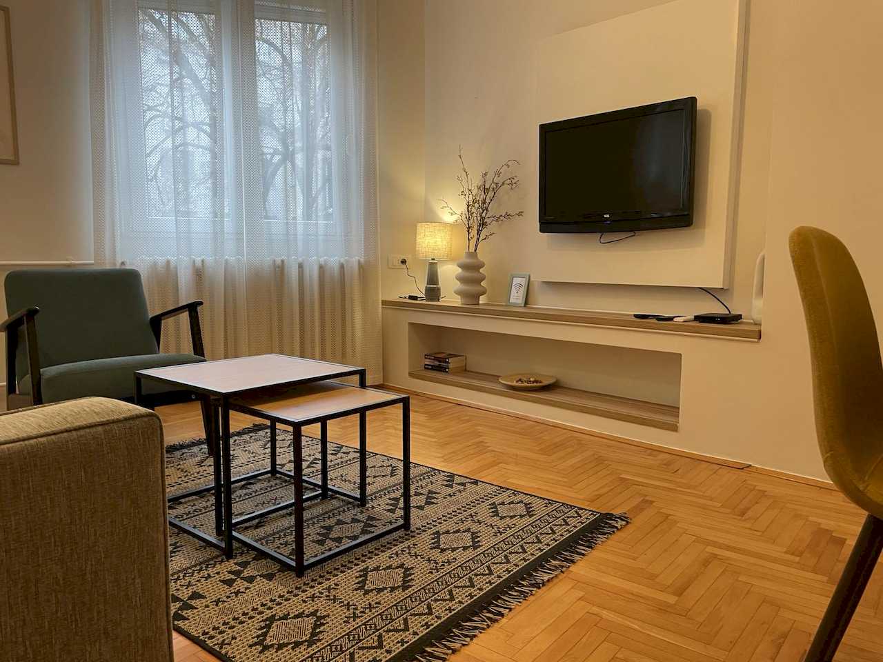 Living area with nesting coffee tables on a geometric rug, TV on white panel, teal armchair, partial grey sofa and window with white curtains – Klishe Zen Apartment – City Break Apartments
