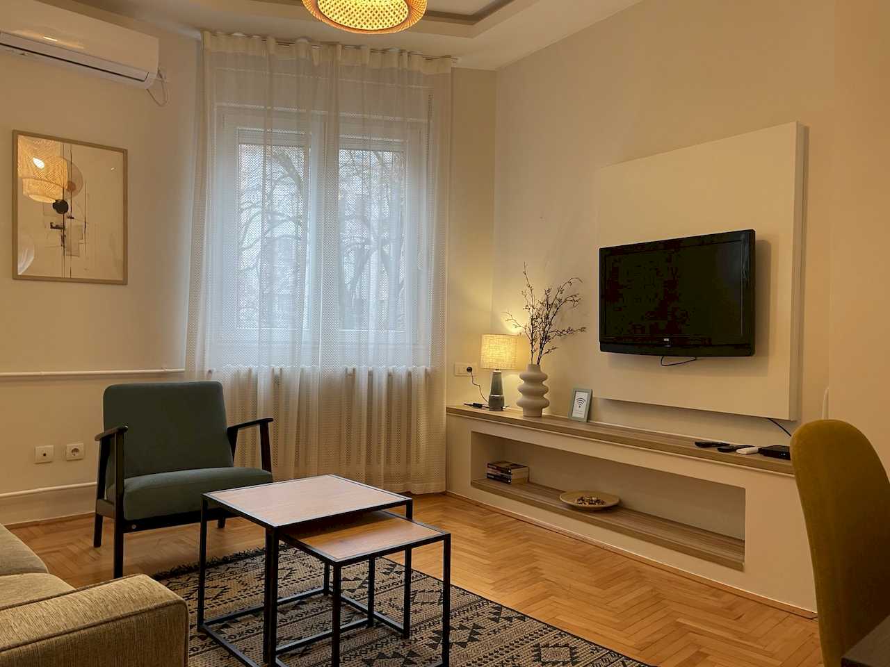 Living room with TV, teal armchair by the window, nesting tables, geometric rug, woven pendant, framed artwork and wall air conditioner – Klishe Zen Apartment – City Break Apartments