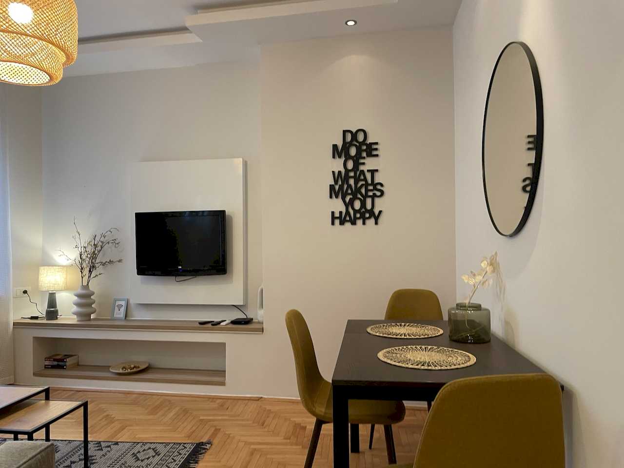Dining area with black table and mustard chairs, woven placemats, round mirror, wall quote and TV zone in the background – Klishe Zen Apartment – City Break Apartments