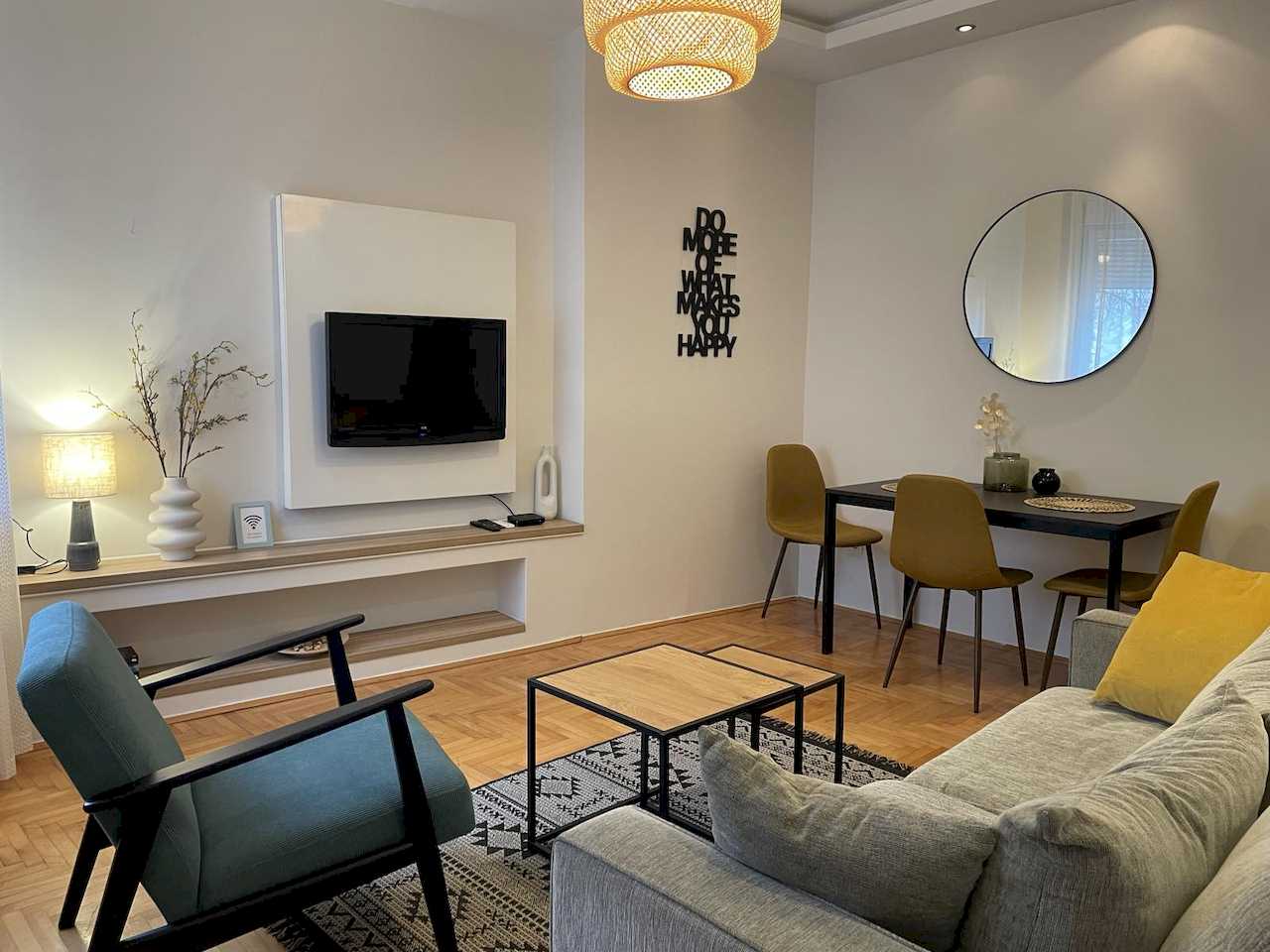 View across living and dining – grey sofa, yellow cushion, teal armchair, nesting tables, black table with mustard chairs, round mirror, woven pendant and wall quote – Klishe Zen Apartment – City Break Apartments