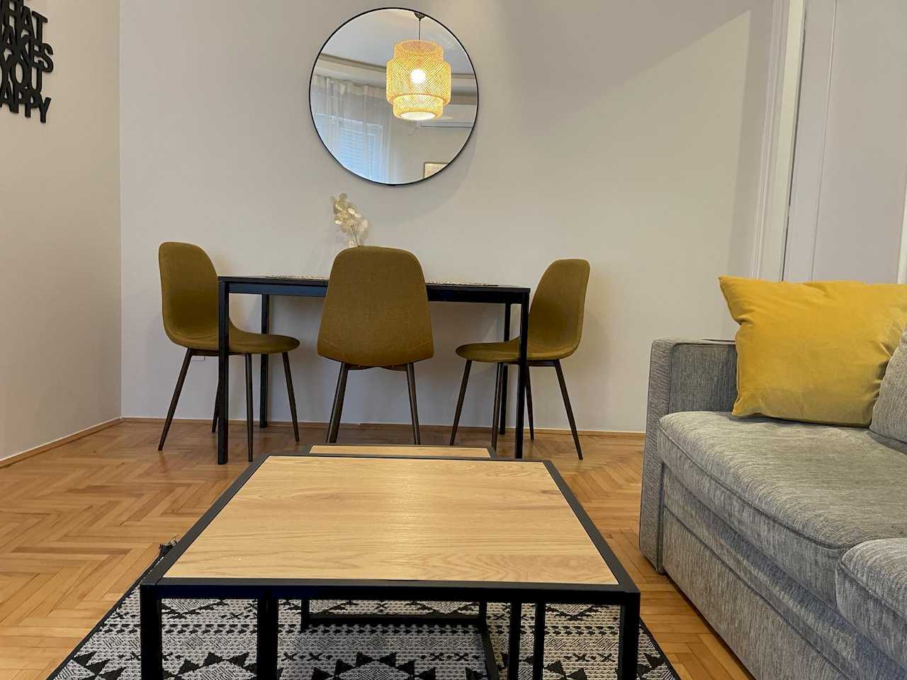 Dining area with three mustard chairs and round mirror, partial grey sofa with yellow cushion and wood coffee table – Klishe Zen Apartment – City Break Apartments