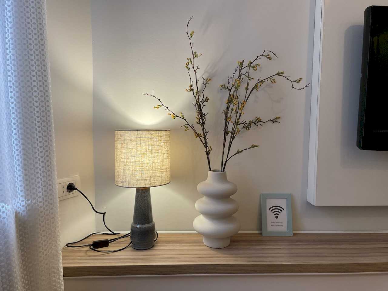 TV shelf detail – table lamp, white vase with dried branches and light-blue Wi-Fi frame – Klishe Zen Apartment – City Break Apartments