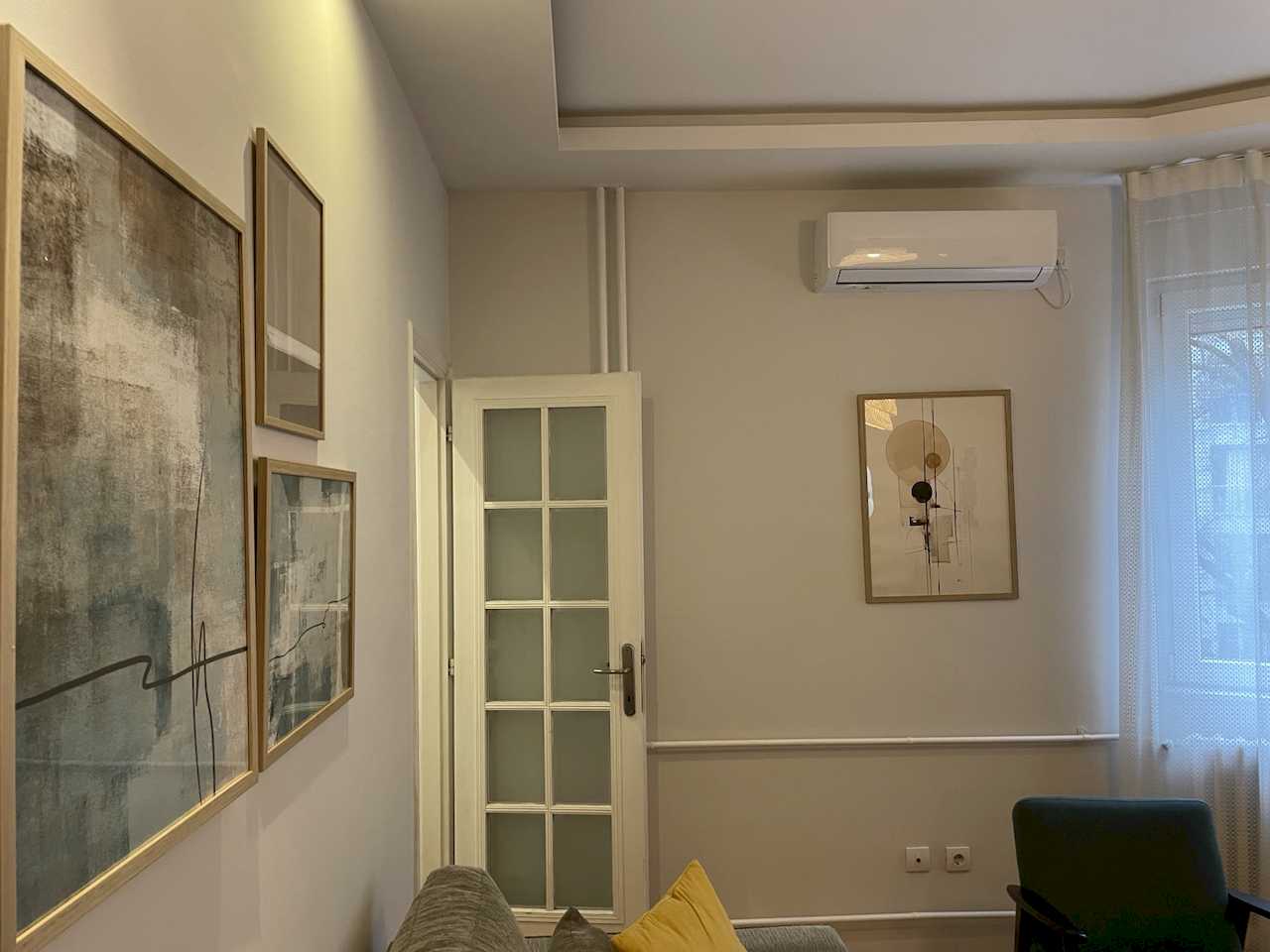 Living area with abstract gallery wall, white glazed panel door, air conditioner and sofa corner with yellow cushion – Klishe Zen Apartment – City Break Apartments