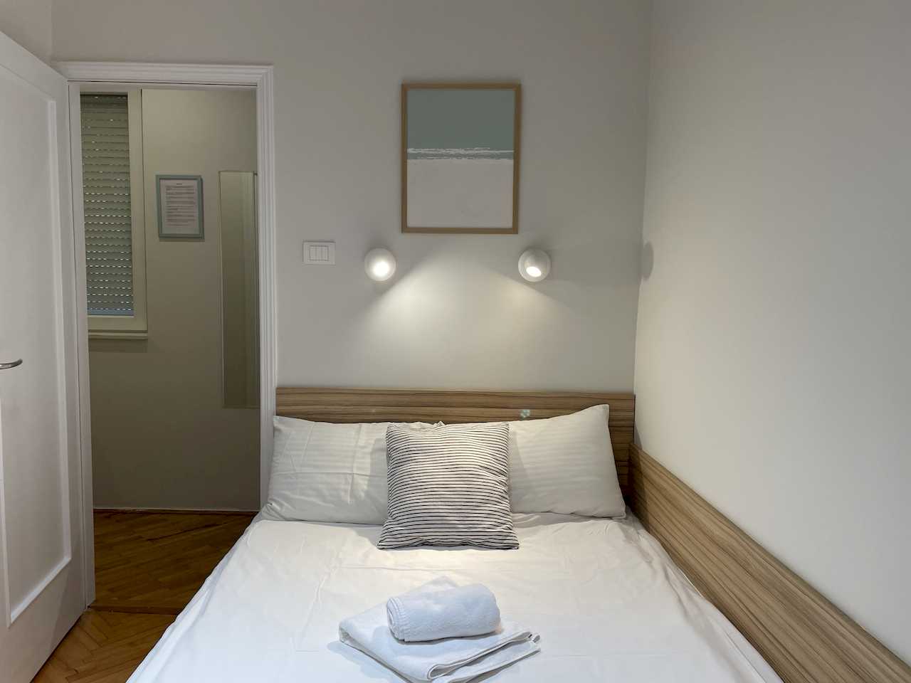 Bedroom with double bed, light wood headboard, white linens, rolled towels, abstract art and lit round wall reading lights – Klishe Zen Apartment – City Break Apartments