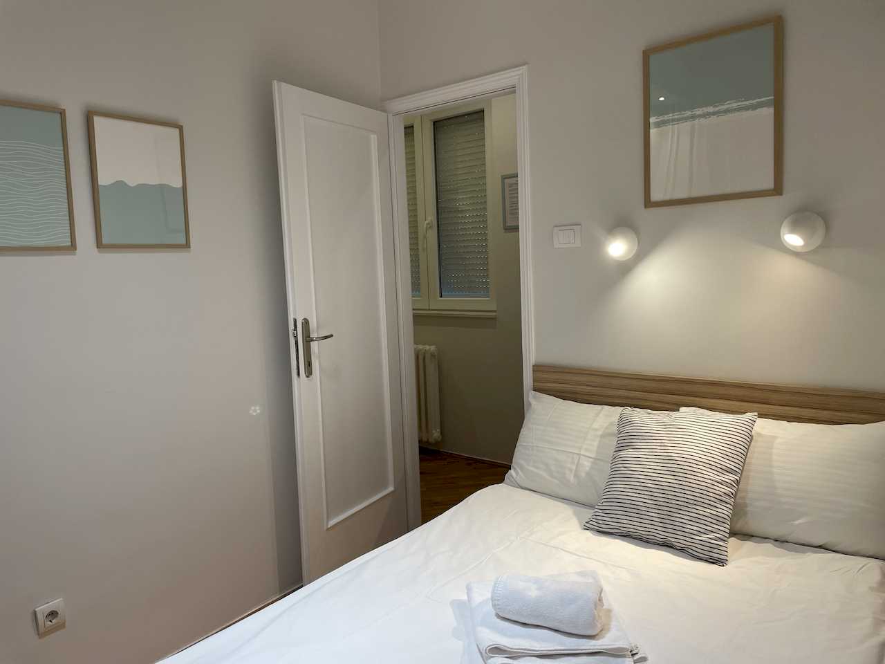 Bedroom with double bed, wood headboard, wall reading lights, framed artwork and open door to the adjoining room – Klishe Zen Apartment – City Break Apartments
