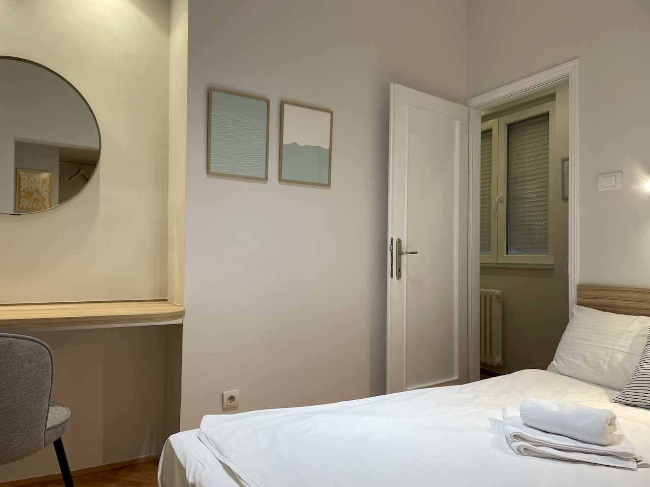 Bedroom with bed, floating wood desk and large round mirror, grey chair and abstract pictures – Klishe Zen Apartment – City Break Apartments