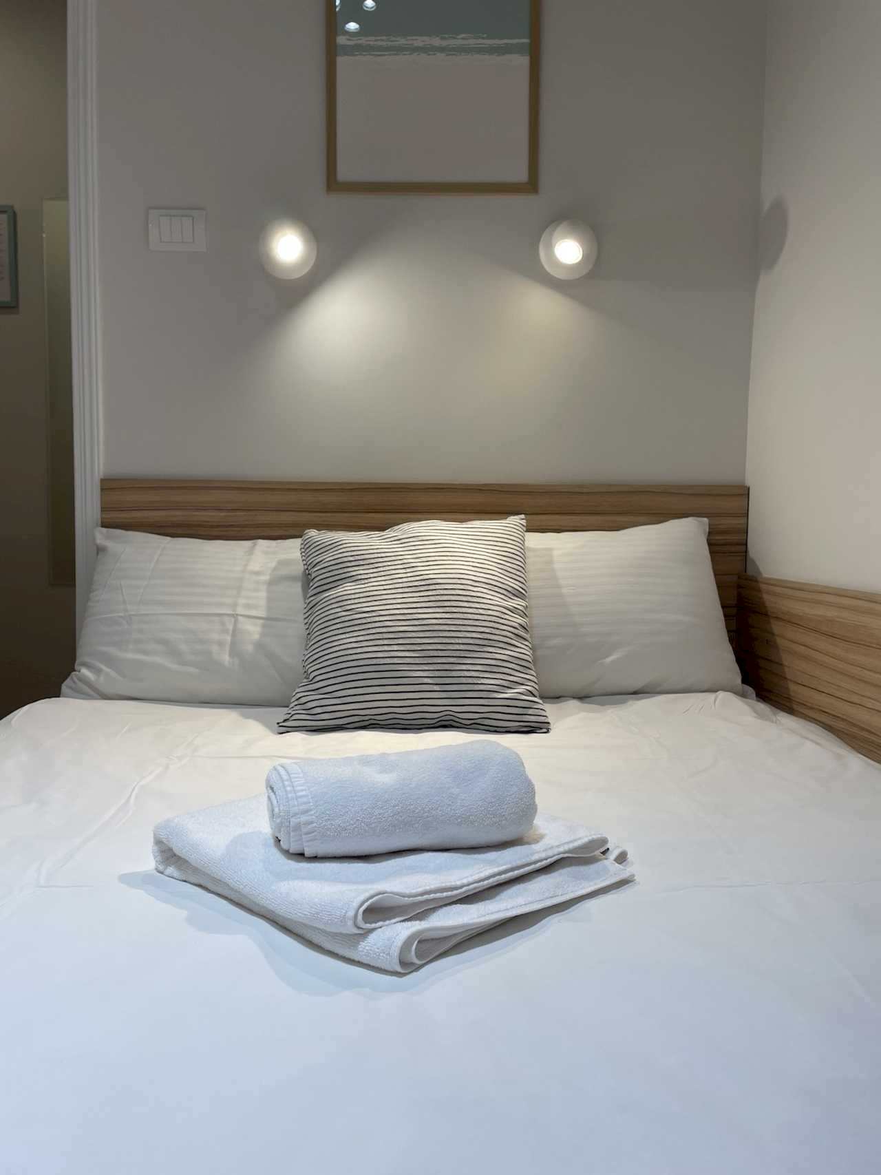 Close-up of double bed with white linens, striped accent pillow, rolled towels, wood headboard and lit wall-mounted reading lights – Klishe Zen Apartment – City Break Apartments