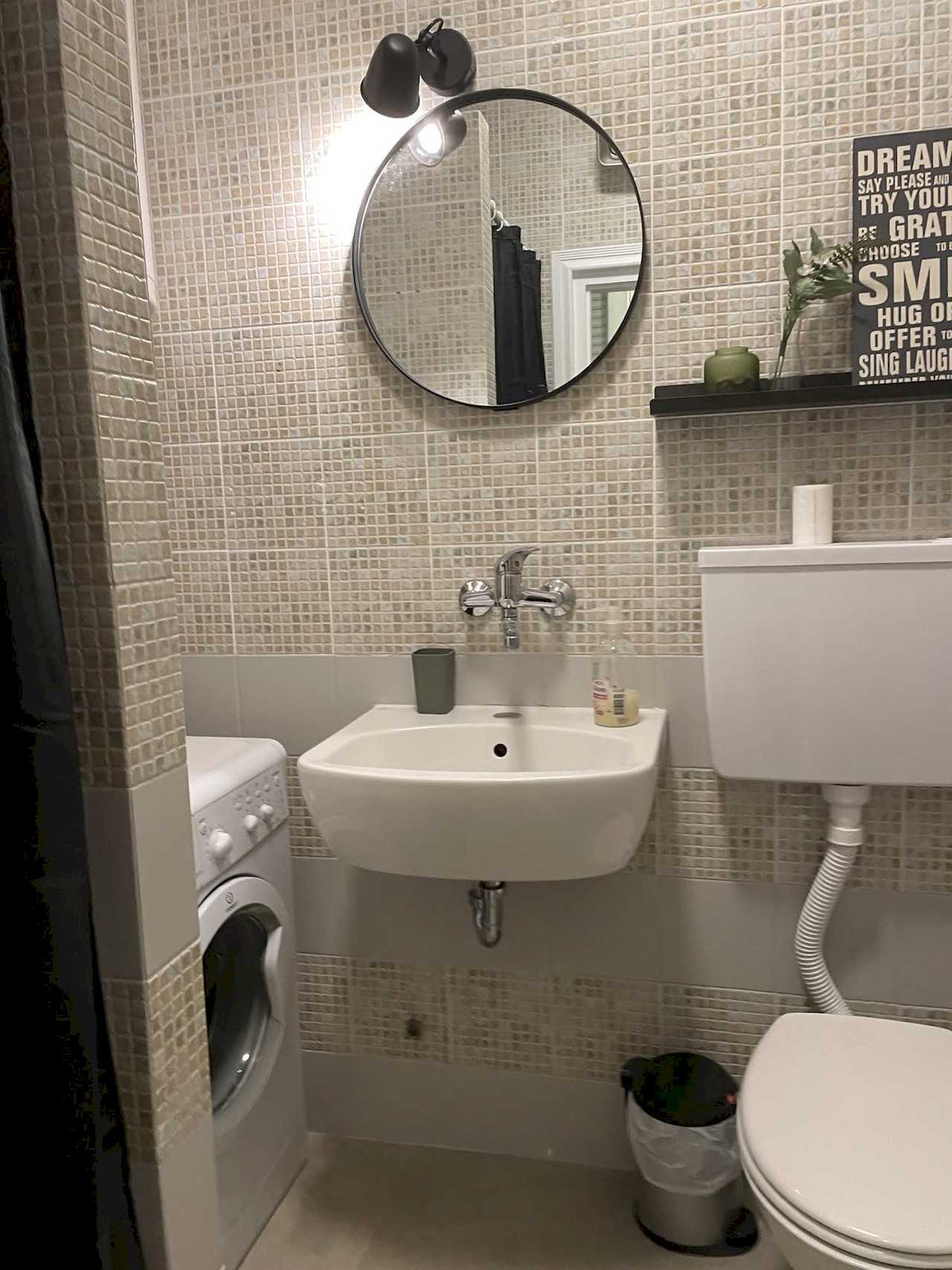 Bathroom with mosaic tiles, white wall-mounted sink, round mirror, black two-light fixture, washing machine and toilet – Klishe Zen Apartment – City Break Apartments
