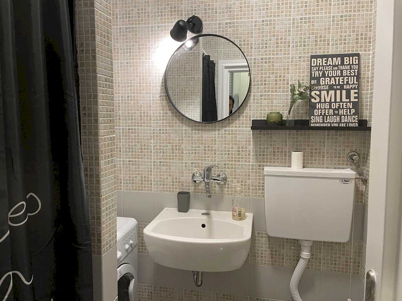 Bathroom with mosaic walls, wall sink, round mirror, white toilet tank and black shelf with letter board decor – Klishe Zen Apartment – City Break Apartments