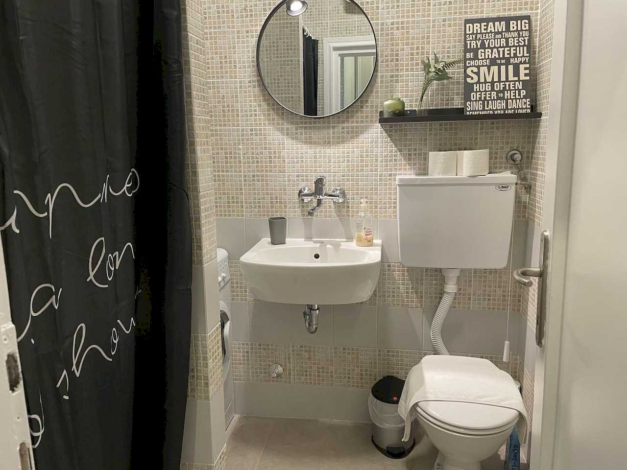 Bathroom with white sink and toilet, mosaic tiles, black shower curtain with white lettering, washing machine and decorative shelf – Klishe Zen Apartment – City Break Apartments