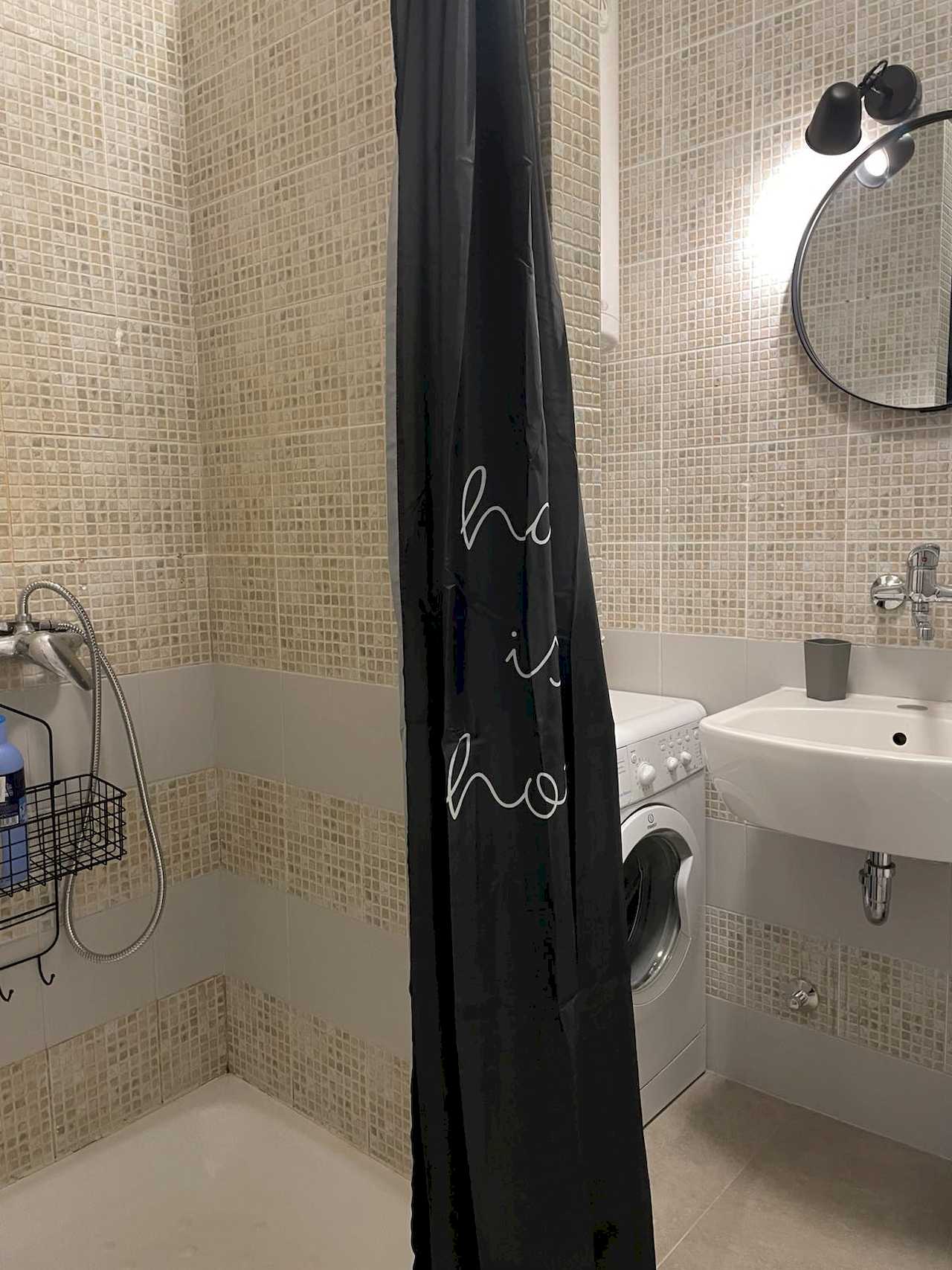 Bathroom – shower with chrome fittings and black curtain, white washing machine, wall sink with round mirror and mid-height band of larger grey tiles – Klishe Zen Apartment – City Break Apartments