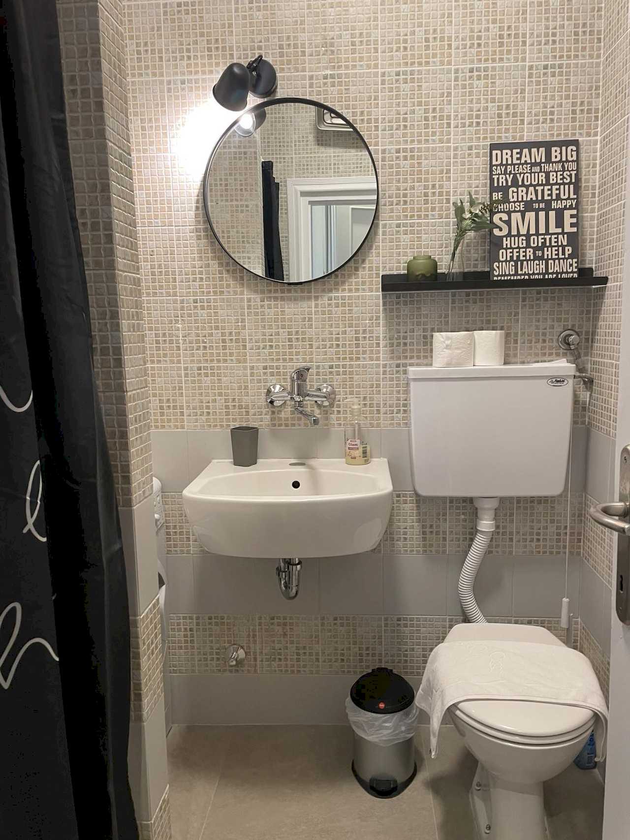 Bathroom with mosaic tiles, white sink and toilet, round mirror, black shelf with letter board and partial shower curtain – Klishe Zen Apartment – City Break Apartments