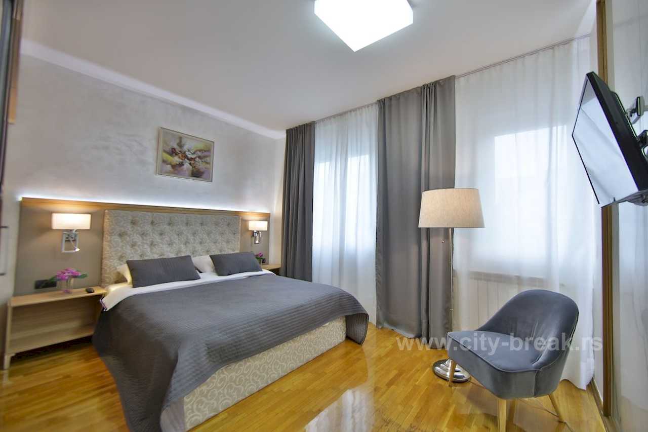 Spavaća soba sa bračnim krevetom, sivom foteljom, LED policom iznad kreveta i TV-om – Apartman Merkator – City Break Apartments