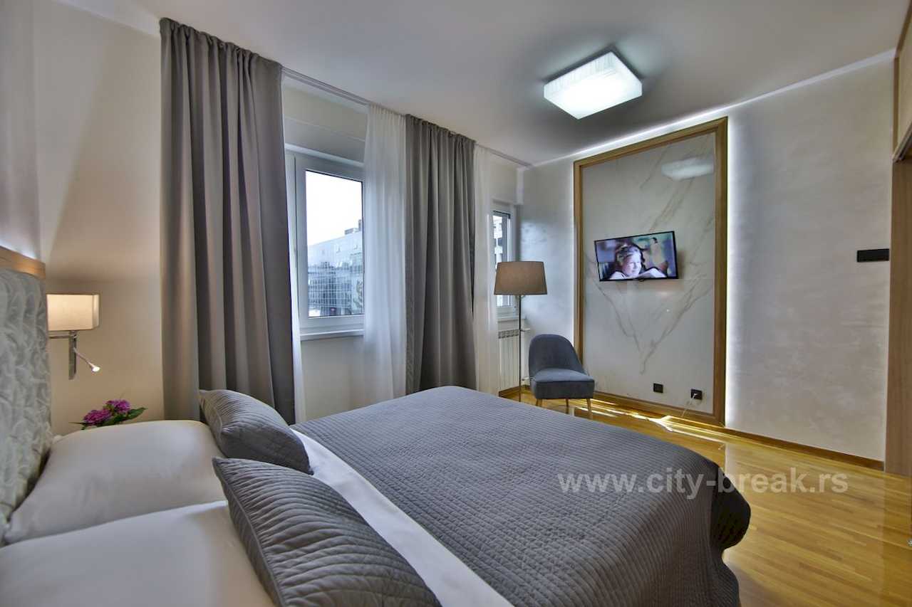 Spavaća soba sa bračnim krevetom, mermernim zidom iza TV-a i foteljom pored prozora – Apartman Merkator – City Break Apartments