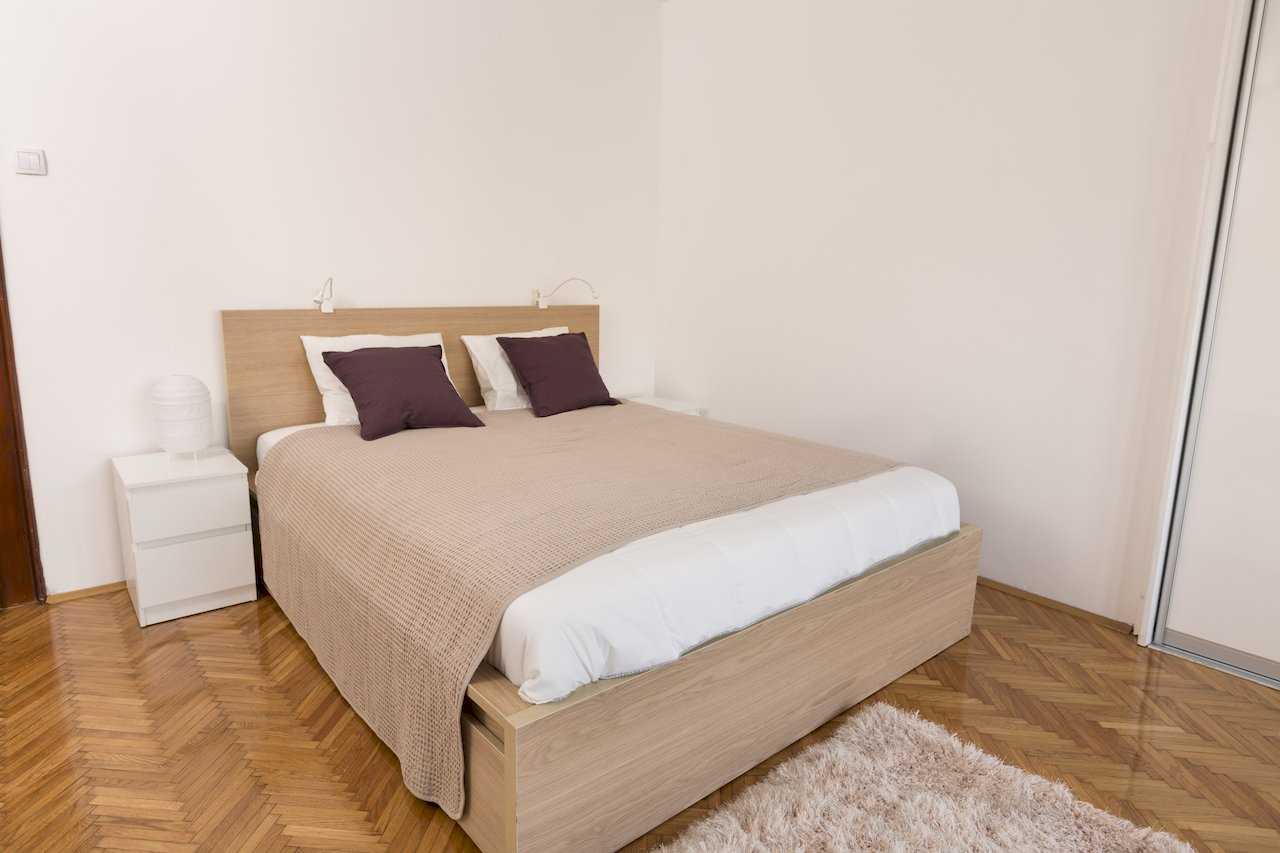 Bračni krevet sa svetlim drvenim okvirom, belim noćnim ormarićem i ljubičastim jastucima – Apartman Milica – City Break Apartments