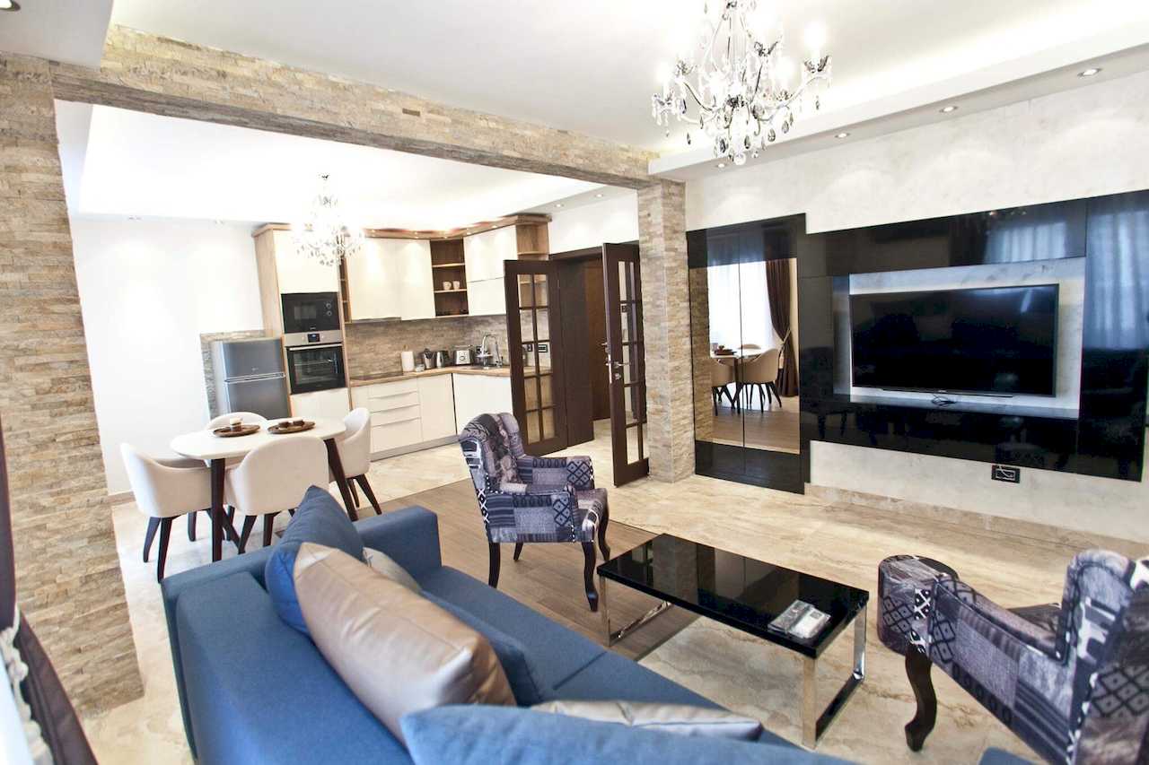 Dnevna soba sa TV-om i ogledalom, u pozadini ovalni trpezarijski sto i kuhinja odeljeni kamenom – Apartman Molerova – City Break Apartments