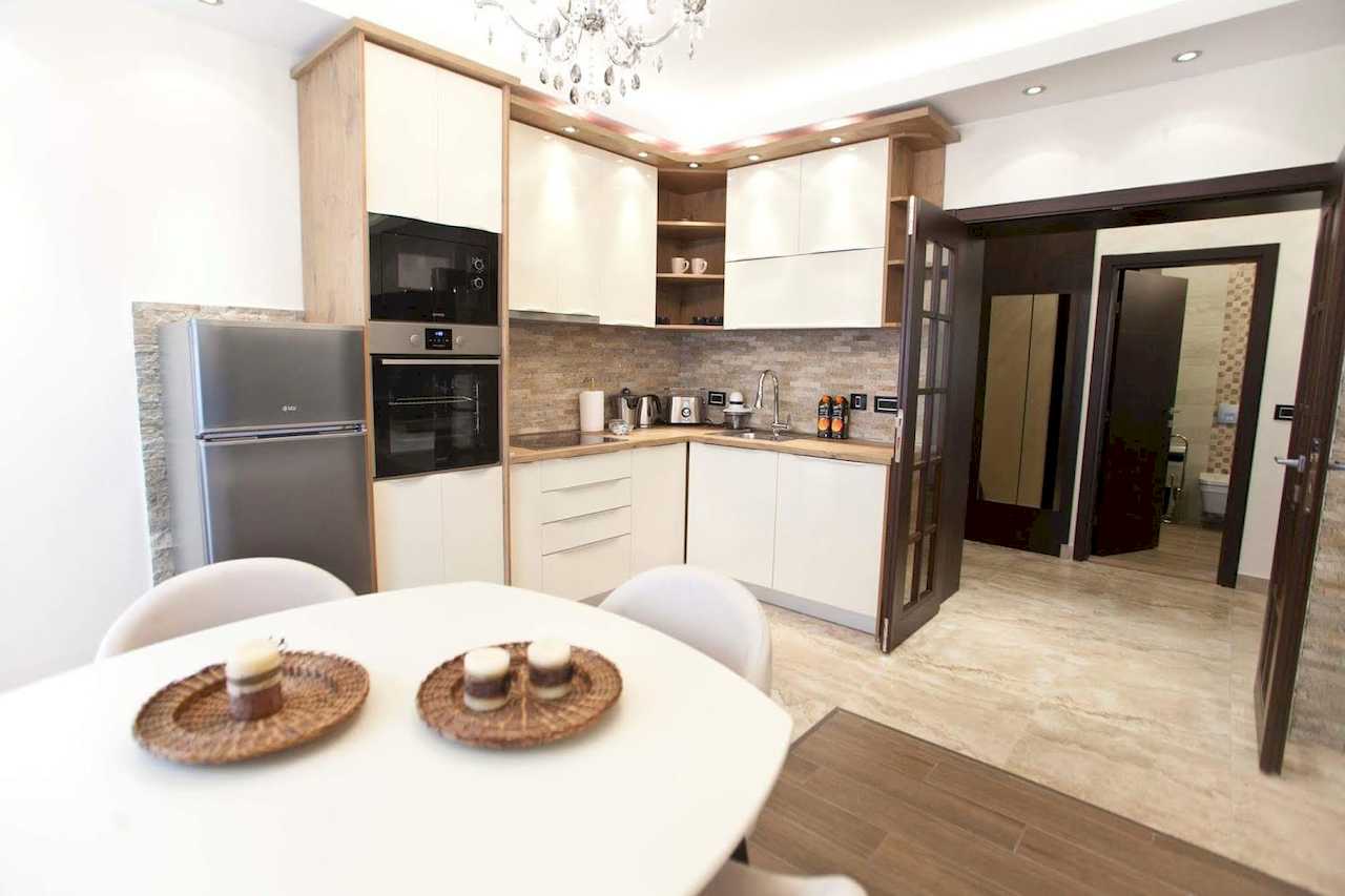 Kuhinja u L-formi sa belim elementima, kamenom na zidu i okruglim trpezarijskim stolom – Apartman Molerova – City Break Apartments