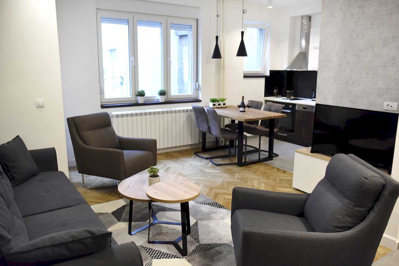 Dnevna soba sa sofom i foteljama, trpezarija i kuhinja u pozadini, parket riblja kost – Apartman Mondo 2 – City Break Apartments