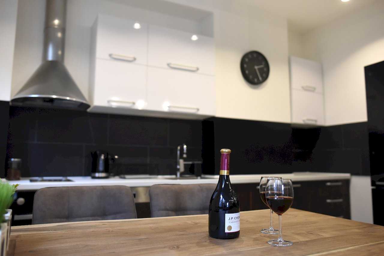 Trpezarijski sto sa vinom u prvom planu i kuhinja sa crno-belim pločicama – Apartman Mondo 2 – City Break Apartments