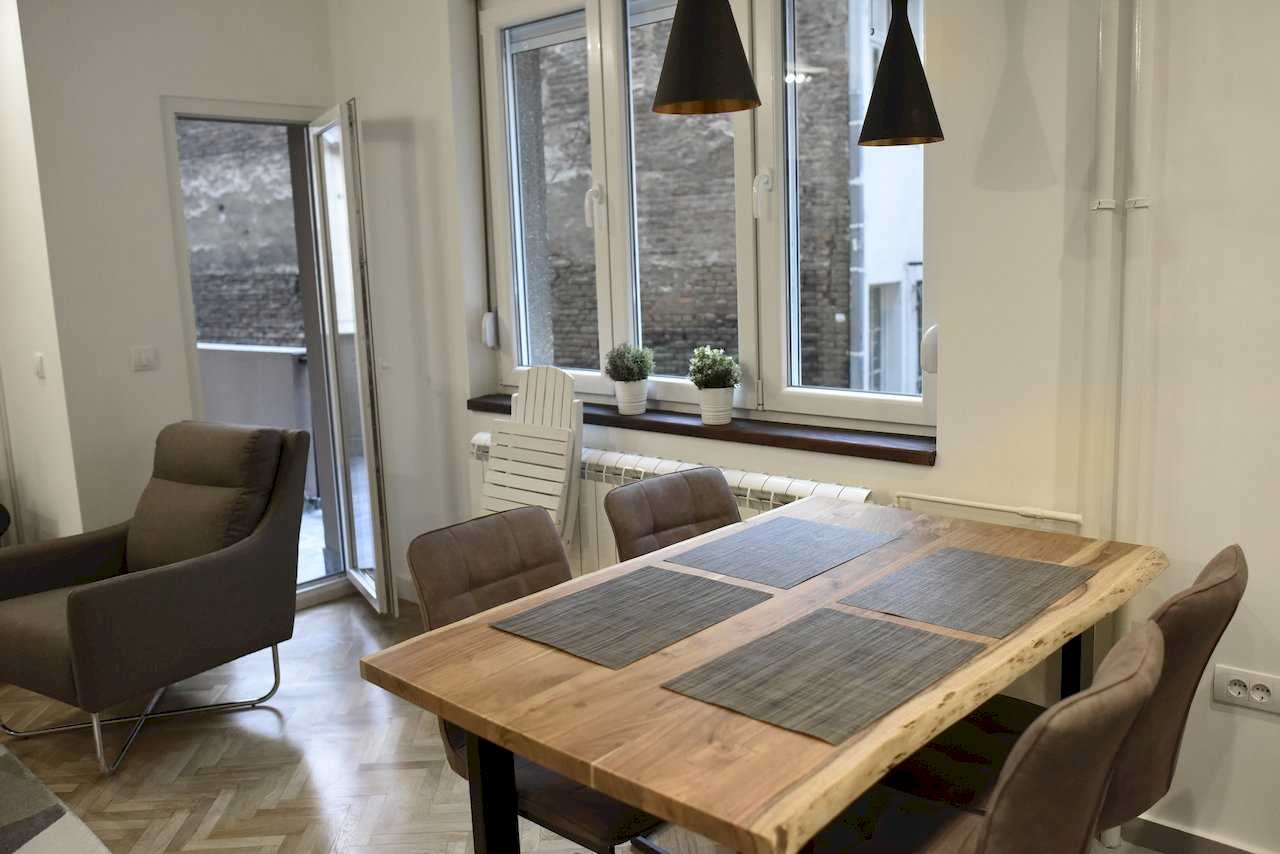 Trpezarija uz prozor i otvorena vrata ka terasi, drveni sto i braon stolice – Apartman Mondo 2 – City Break Apartments