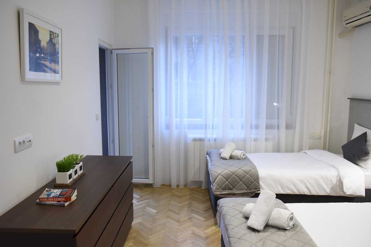 Spavaća soba sa dva pojedinačna kreveta, tamna komoda i prozor sa belim zavesama – Apartman Mondo 2 – City Break Apartments