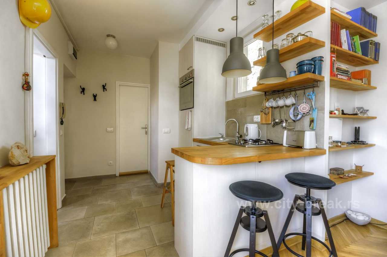 Apartman NINA (Vračar, Beograd) – kuhinja sa belim šankom, drvenom radnom pločom, crnim barskim stolicama i otvorenim policama