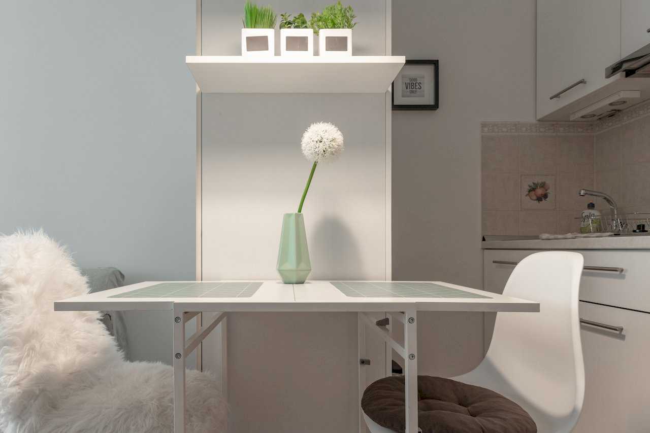 Dining table with white chairs, plants on a shelf above, and kitchen in the background – Apartment Oaza – City Break Apartments