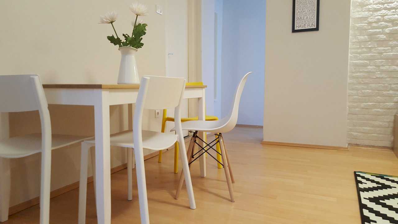 Light-wood dining table with white and yellow chairs, white floral centerpiece, and partial white brick wall – Apartment Opera – City Break Apartments