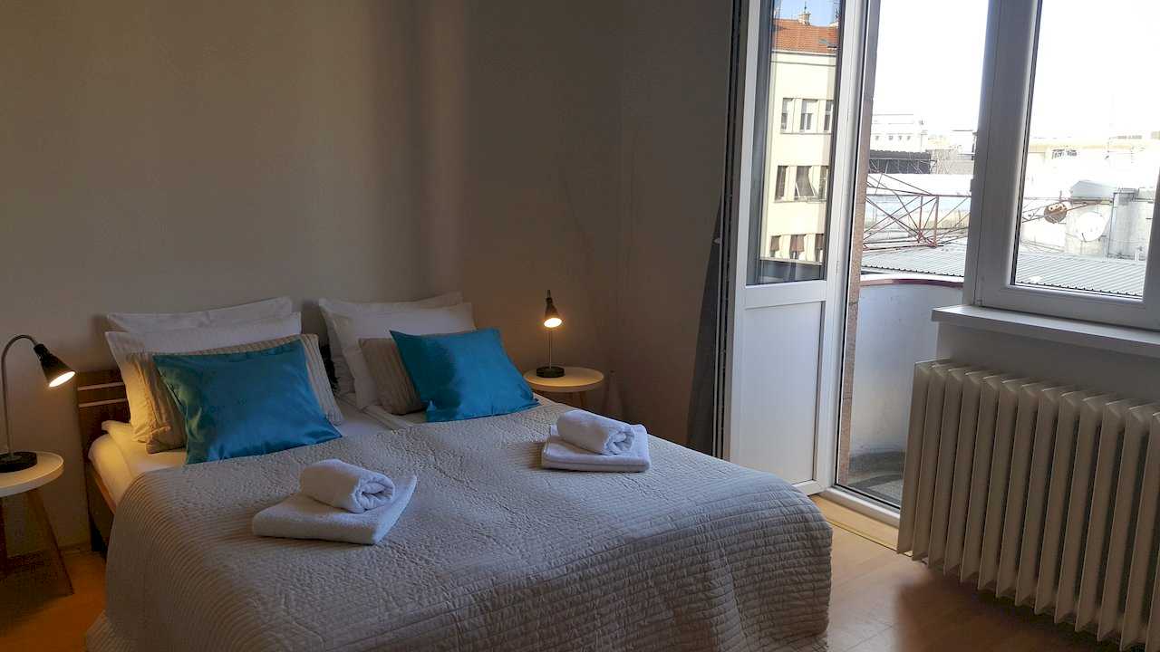 Bedroom with double bed, turquoise pillows, rolled white towels, and glass door to balcony overlooking city buildings – Apartment Opera – City Break Apartments