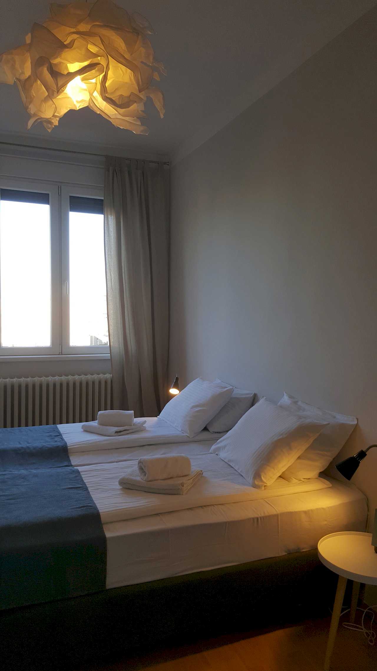 Bedroom with two joined single beds, white linens, navy throw at the foot, rolled towels, curtains, and sculptural pendant light – Apartment Opera – City Break Apartments