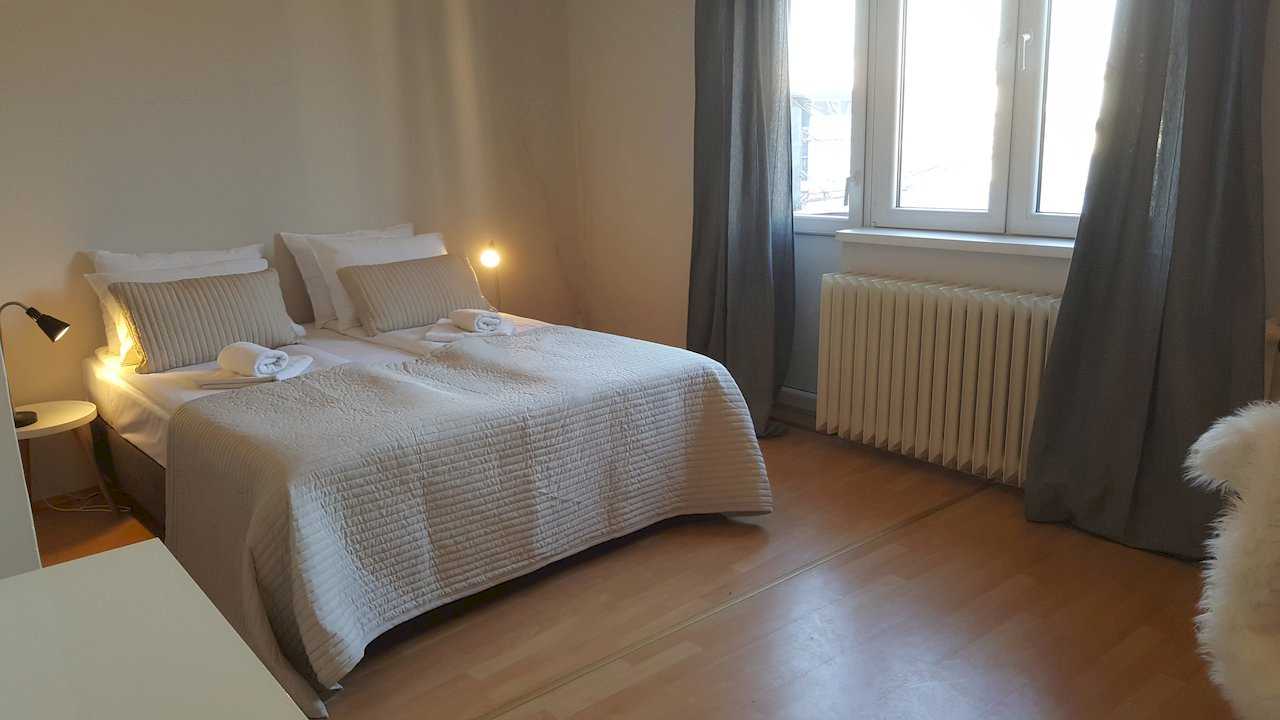 Bedroom with beige quilted bedspread, cushions, rolled towels, large window with dark curtains, and bedside lamps – Apartment Opera – City Break Apartments