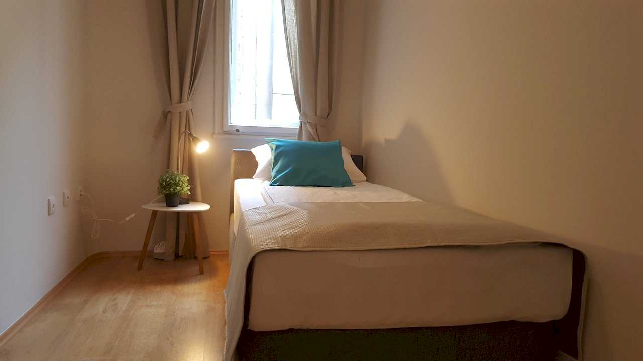 Single bed with turquoise pillow, round white nightstand with lit black desk lamp and small plant – Apartment Opera – City Break Apartments