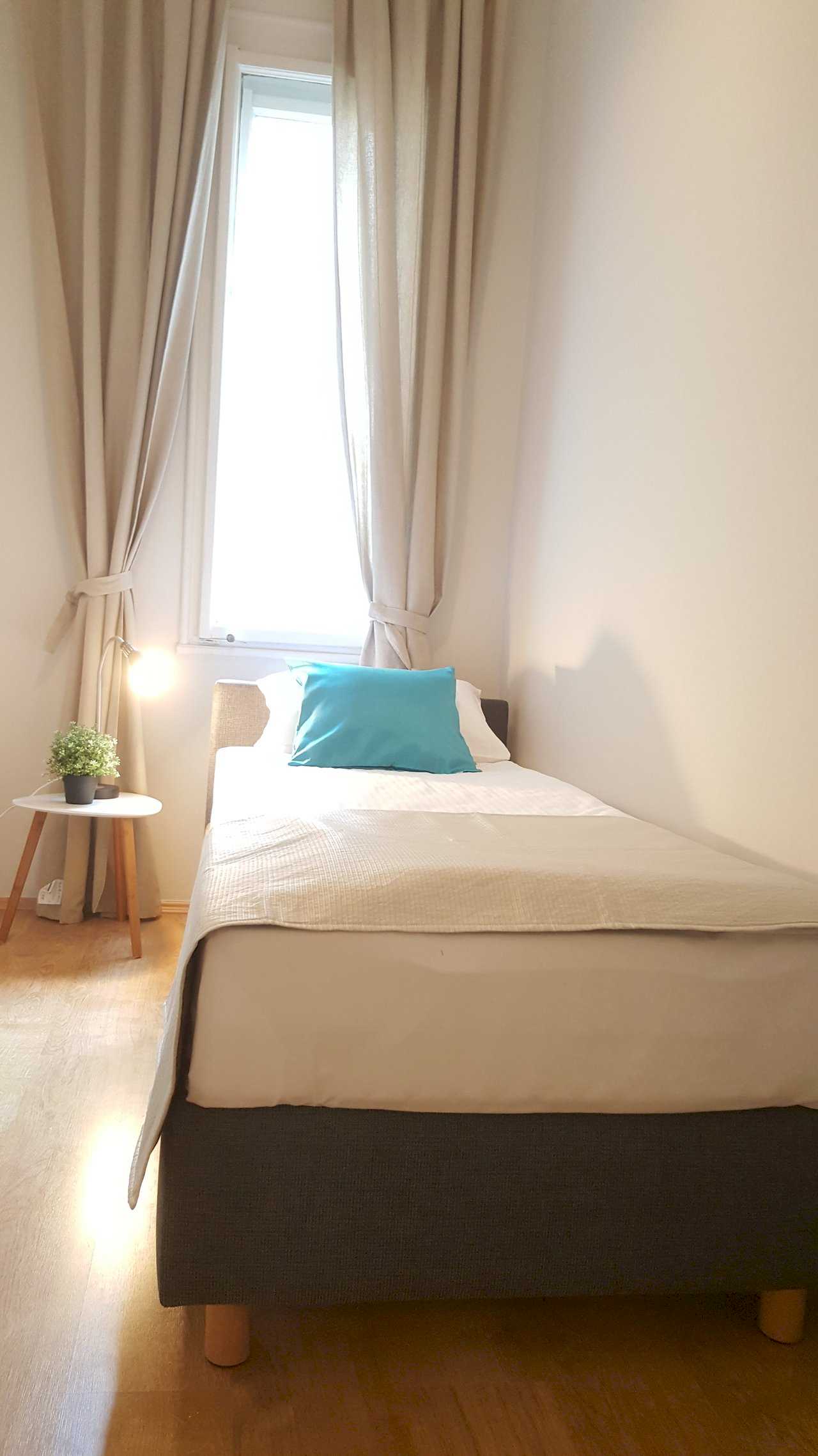 Single bed with grey upholstered frame, beige throw, turquoise accent pillow, and tall window with tied-back beige curtains – Apartment Opera – City Break Apartments