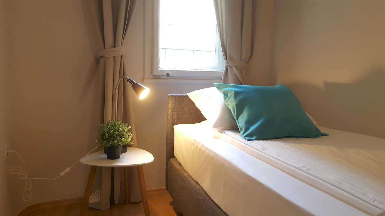 Single bed beside window with tie-back beige curtains, triangular white nightstand, lamp, and plant – Apartment Opera – City Break Apartments
